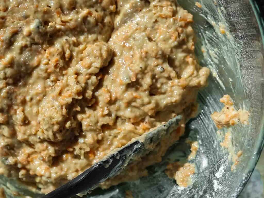 Close-up of a bowl containing thick, light brown vegan carrot cake batter with small visible bits of grated carrot. A mixing spoon is partially visible, coated with the batter. The glass bowl shows some flour residue on its side.